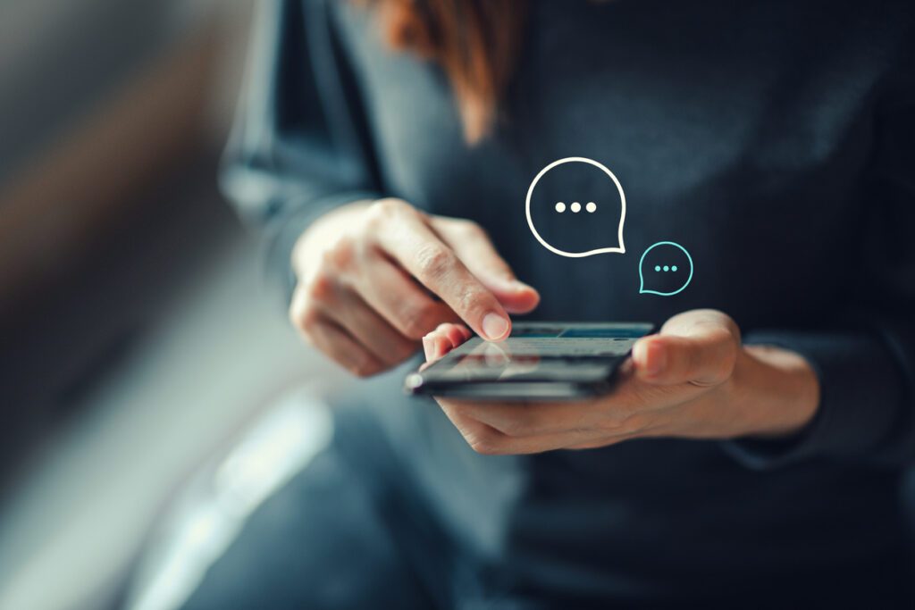 Woman’s hand typing on a smartphone with text bubbles to represent one form of consumer messaging via SMS.